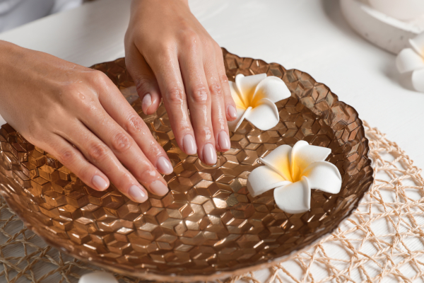 Woman soaking her hands in bowl with water and flowers on table, closeup. Spa treatment